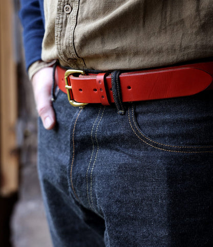 A person wearing a tan button-up shirt, dark blue jeans, and the Best Made Co. Famous Red Belt—a bold red leather belt with a gold buckle made in the USA—with one hand in their pocket.