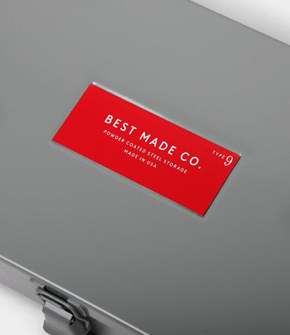 A close-up of the Best Made Co. Type 9 Strongbox, a durable gray steel storage box with a red label and white text, featuring powder-coated steel and a sturdy bottom latch for secure storage. Made in USA.