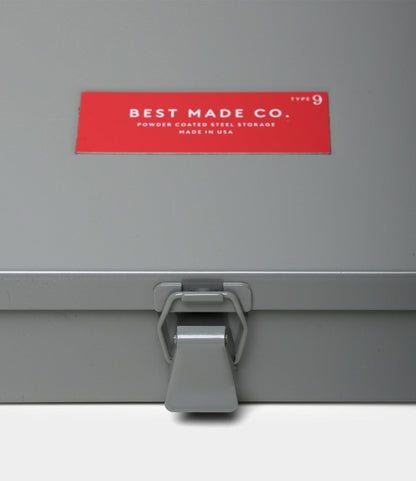 A close-up of a gray Type 9 Strongbox by Best Made Co., featuring a latch and a red label that reads: "BEST MADE CO. Type 9, Powder Coated Steel Storage, Made in USA.