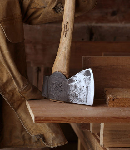 A close-up of The Phantom, a hand-forged felling axe by Best Made Co., with a wooden handle on stacked boards in a workshop. A brown work jacket hangs nearby against a backdrop of wooden surfaces.