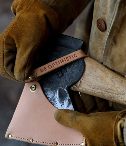A person in brown gloves and a tan jacket holds The Phantom felling axe by Best Made Co., featuring a wooden handle and a leather cover embossed with "BE OPTIMISTIC.