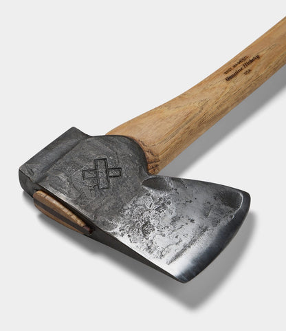 A close-up of Best Made Co.’s The Phantom felling axe, showing its hand-forged, worn high-carbon steel blade with an engraved cross symbol and a light wooden handle with text. The background is plain white.