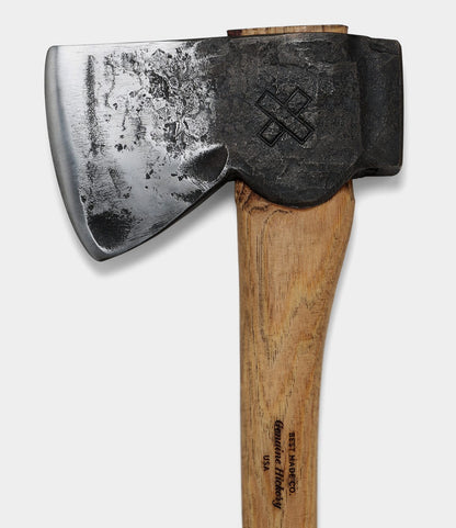 A close-up of The Phantom by Best Made Co.—a hand-forged felling axe with a polished high-carbon steel blade, weathered head engraved with an X, and a wooden handle stamped with text and showing visible grain and wear.