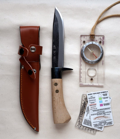 The Hotta San Hunting Knife by Best Made Co., featuring a wooden handle, handmade blade, and brown leather sheath, is displayed with a map compass and map symbol cards on a beige surface.