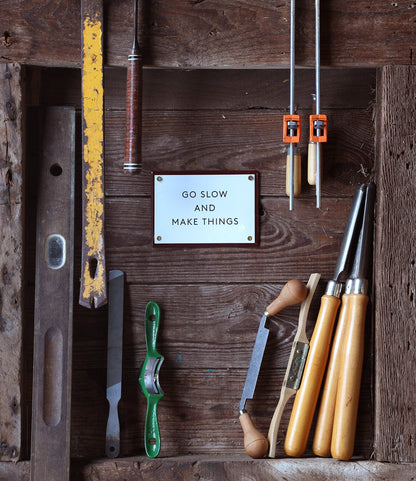 Woodworking tools surround the “Go Slow” Enamel Steel Sign by Best Made Co. Hanging this durable wall decor adds charm to your workshop and complements your clamps, chisels, and spokeshave.