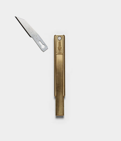 A brass Best Made Co. Studio Knife with a rectangular handle lies next to its detached, pointed silver workshop blade on a plain white background.