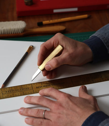 Using a Best Made Co. Studio Knife and a metal ruler, a person in a denim shirt with a wedding ring cuts white material; a pencil, brush, and other tools are on the table.