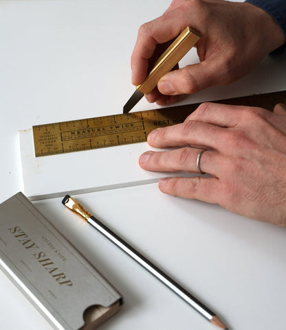 A person uses a brass ruler and a Best Made Co. Studio Knife to score a white board. Nearby, a pencil and a Stay Sharp metal box rest on the table, while the person wears a simple wedding ring.