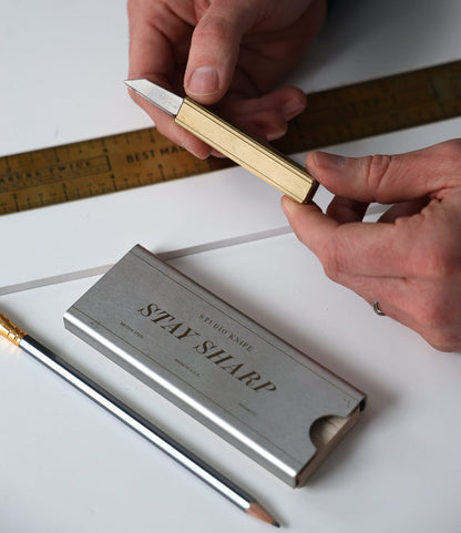 A person holds a Best Made Co. Studio Knife above a ruler, with a pencil and a metal case labeled "STAY SHARP" on a white surface.