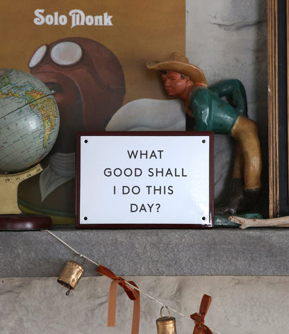 A shelf displays a small globe, a forward-leaning cowboy figurine, a “Solo Monk” album cover, and Best Made Co.’s “What Good” Enamel Steel Sign. Beneath the shelf hangs a string with bells.