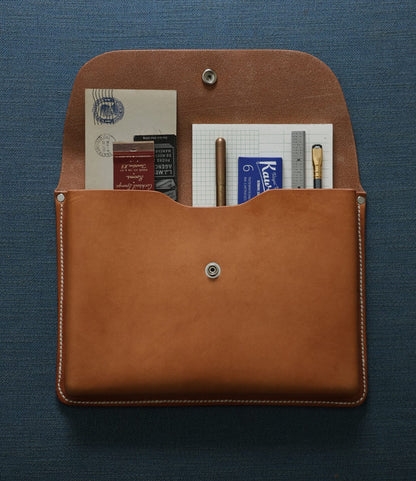 The Best Made Co. Gfeller Case Small, a brown hand-stitched leather case with a snap button, is shown open on blue fabric, displaying notebooks, envelopes, a pen, pencil, and ruler inside.