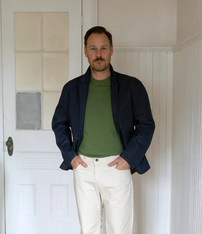 A man stands indoors by a white door, wearing a dark denim jacket, light-colored pants, and the Best Made Co. Standard T-Shirt in Olive Drab, with his hands in his pockets, looking at the camera.