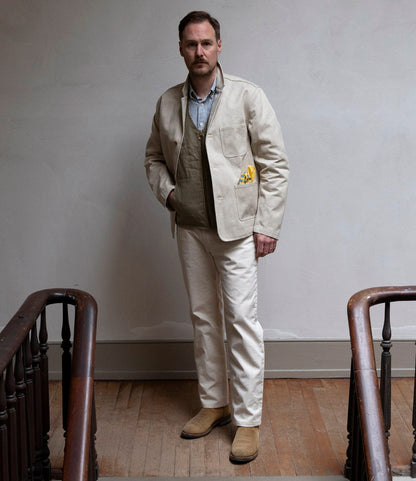 A man stands indoors on a wooden floor between two stair railings, wearing the Best Made Co. Selvedge Natural Studio Jacket with a yellow handkerchief in the pocket, along with American craftsmanship-inspired cream pants and tan boots.