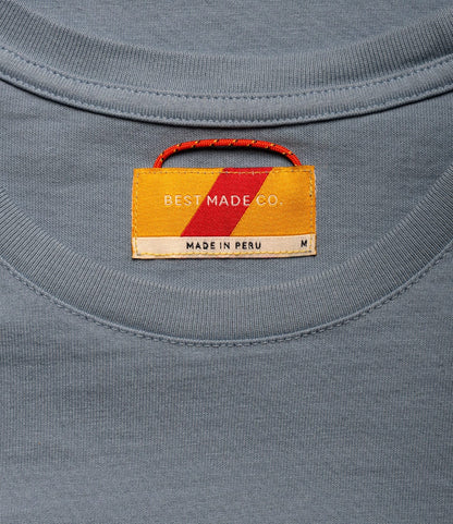 A close-up of the Standard T-Shirt Overcast Blue by Best Made Co. shows a gray garment-dyed neckline with a sewn-in tag, red and yellow stripes, white brand text, size M, and MADE IN PERU—crafted from premium Peruvian tangüis cotton.