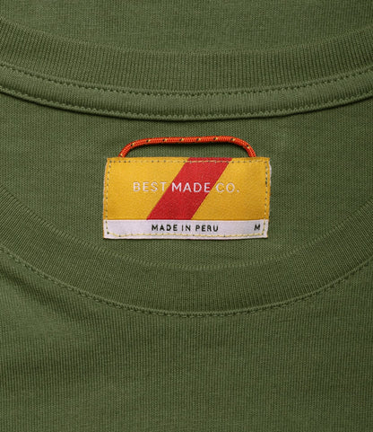 A close-up of the Standard T-Shirt Olive Drab’s collar shows a BEST MADE CO. label with a yellow, red, and white block design—this durable tee is made from Peruvian tangüis cotton, with MADE IN PERU and size M printed beneath.