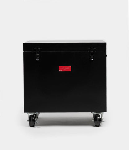 The Best Made Co. Rolling Footlocker is a black rectangular storage trunk made from durable 22-gauge steel, featuring a closed lid, two metal latches, caster wheels, and a red label—shown against a plain light gray background.