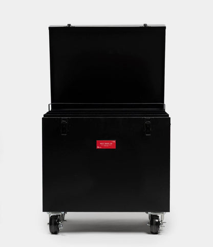 The Best Made Co. Rolling Footlocker is a large, black 22-gauge steel trunk on four caster wheels with its lid open and a small red label on the front, photographed against a plain white background.