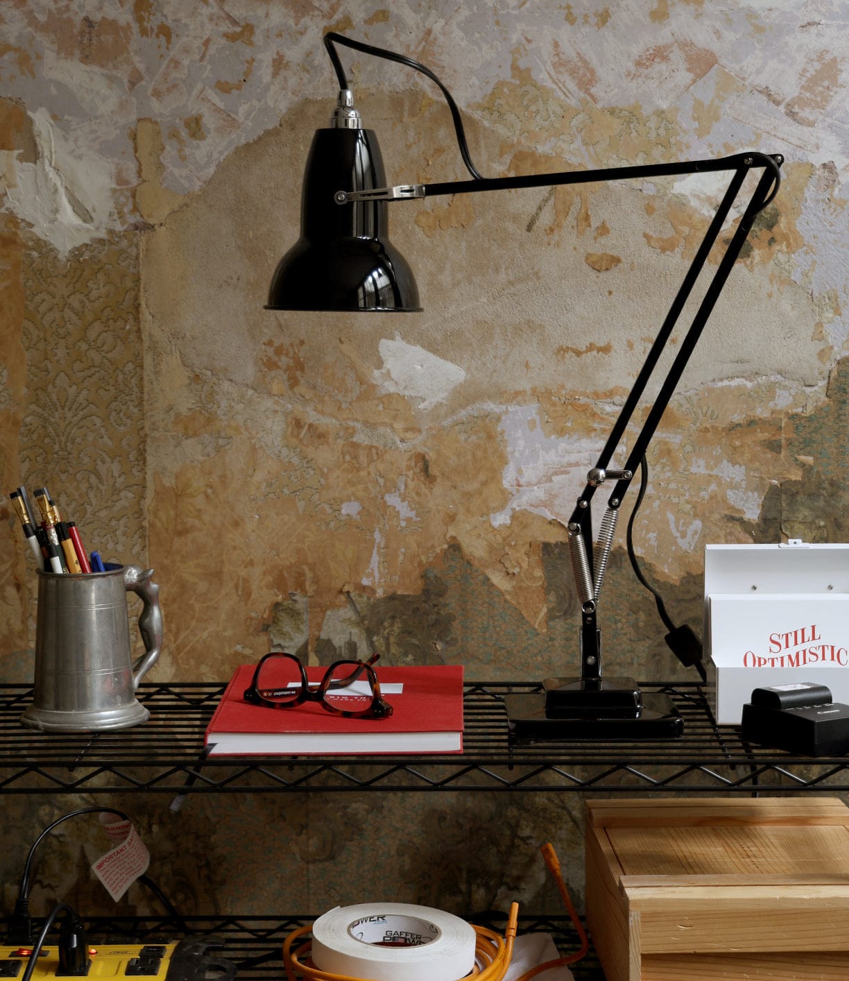 A Best Made Co Anglepoise Original 1227 Desk Lamp sits on a metal shelf beside a mug of pencils, red notebook with glasses, and a sign reading STILL OPTIMISTIC, all set against a distressed, textured wall.