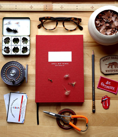 A flat lay of a wooden desk with the Best Made Co. S.B.T. Mid Cloth-Bound Notebook in red, glasses, ruler, pencil, sharpener, fly fishing flies and reel, business cards, scissors, round patch, and “Be Optimistic” and “Honey Badger” embroidered patches.