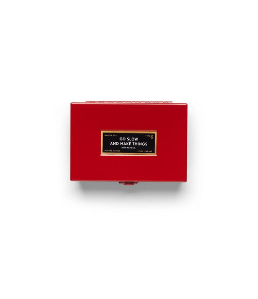 The Best Made Co Type 6 Strongbox, Go Slow Edition, featuring a gold label and clasp, rests on a wooden desk with a pencil and metal sharpener. Above, shelves display yellow and red containers.