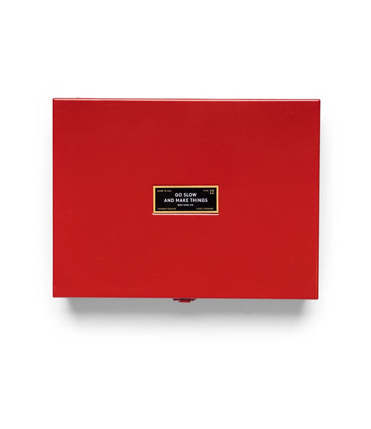A closed, shiny red Best Made Co Type 12 Strongbox, Go Slow Edition with a black label rests on a warm vintage wooden desk beneath cubbyholes filled with small items.