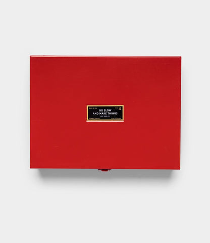 The Type 12 Strongbox, Go Slow Edition by Best Made Co. is a bright red rectangular box featuring a central black label reading "GO SLOW AND MAKE THINGS," shown on a plain light gray background. This storage box is a limited edition item.
