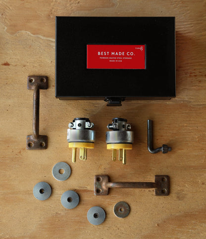 The Type 6 Strongbox - Matte Black by Best Made Co. rests on a wooden surface, accompanied by two industrial handles, two electrical plugs, an L-shaped hex key, and metal washers—perfect for secure, durable storage.