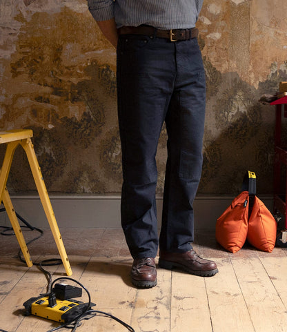 Indoors on a wooden floor, someone wears Best Made Co. Field Pant Special Edition Navy, brown shoes, and a belt; their upper body is partially out of frame. Around them are a yellow tool, cables, yellow sawhorse, and orange bag against a textured wall.