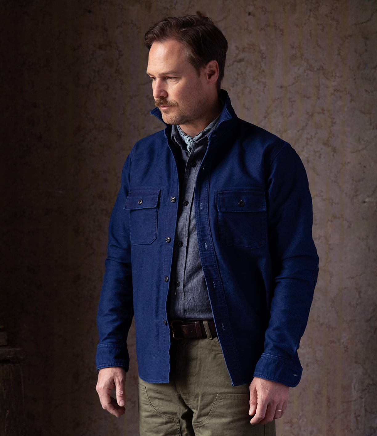 A man with light brown hair and a short beard wears the Best Made Co Indigo Moleskin Work Shirt over a gray tee and olive green pants, standing against a textured brown wall and looking to his left.