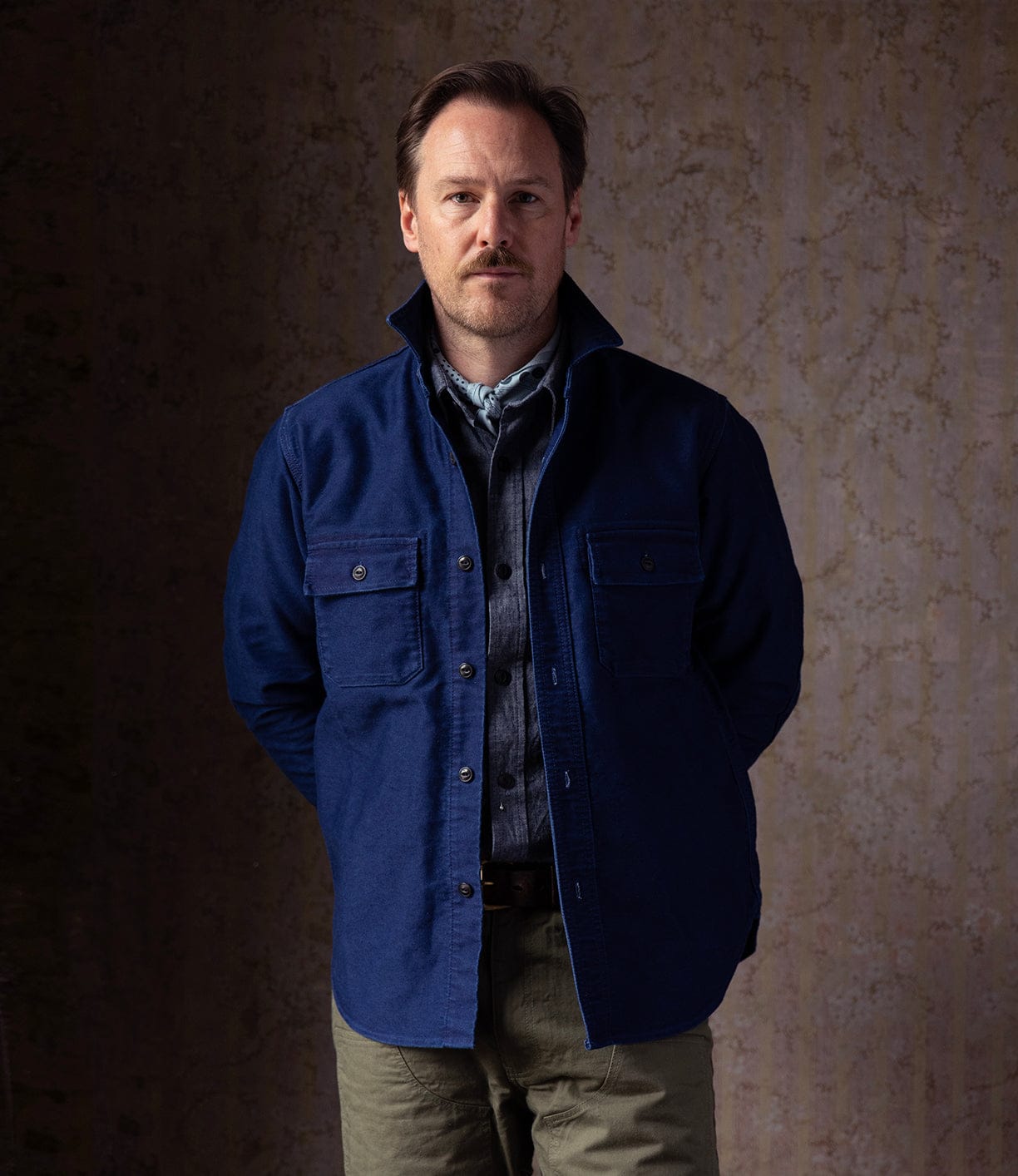 A man with light brown hair and a mustache stands against a textured wall, wearing the Best Made Co Indigo Moleskin Work Shirt under a blue jacket and olive green pants, with his hands behind his back.