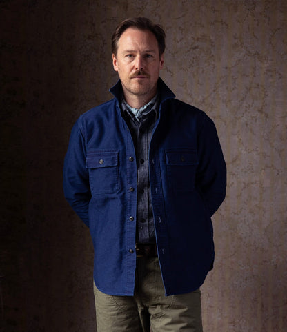 A man with short brown hair and a mustache stands facing the camera, hands behind his back, wearing Best Made Co.'s Indigo Moleskin Work Shirt over a gray shirt and olive pants, set against a neutral, textured background.