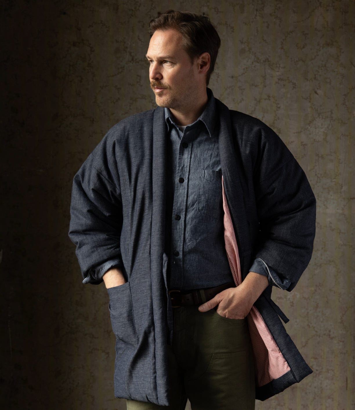 A man in a blue shirt, olive green pants, and the Best Made Co. Quilted Hanten Jacket with red-and-white lining stands with one hand in his pocket, looking to the side against a textured backdrop.