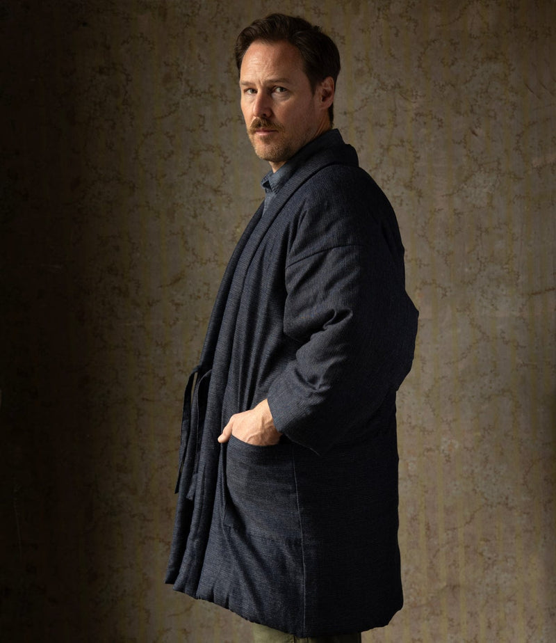 A man with light brown hair and a mustache stands sideways, wearing a dark blue, loose-fitting coat with his hand in the pocket, looking at the camera against a textured, brownish background.