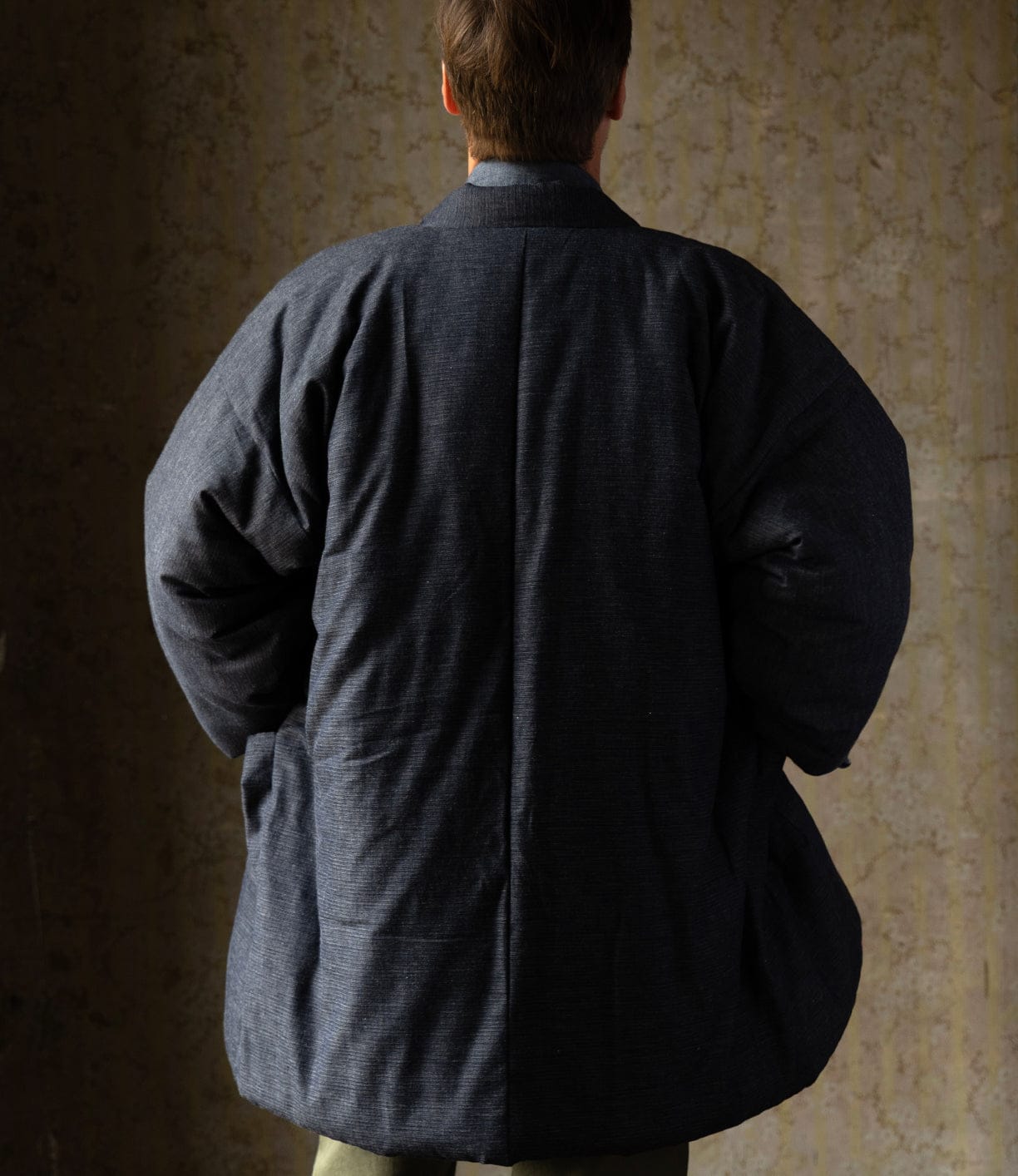 A person faces away from the camera against a textured beige wall, wearing Best Made Co.’s Quilted Hanten Jacket. The oversized, puffy silhouette and Japanese workwear-inspired design are highlighted by their bent arms.