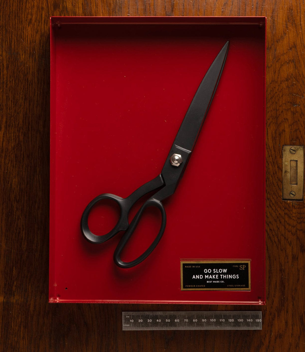 A pair of large, black Kevlar Shears by Best Made Co. is displayed in a red shadow box on a wooden surface, with a small plaque reading "Go slow and make things" and a ruler placed below the box.