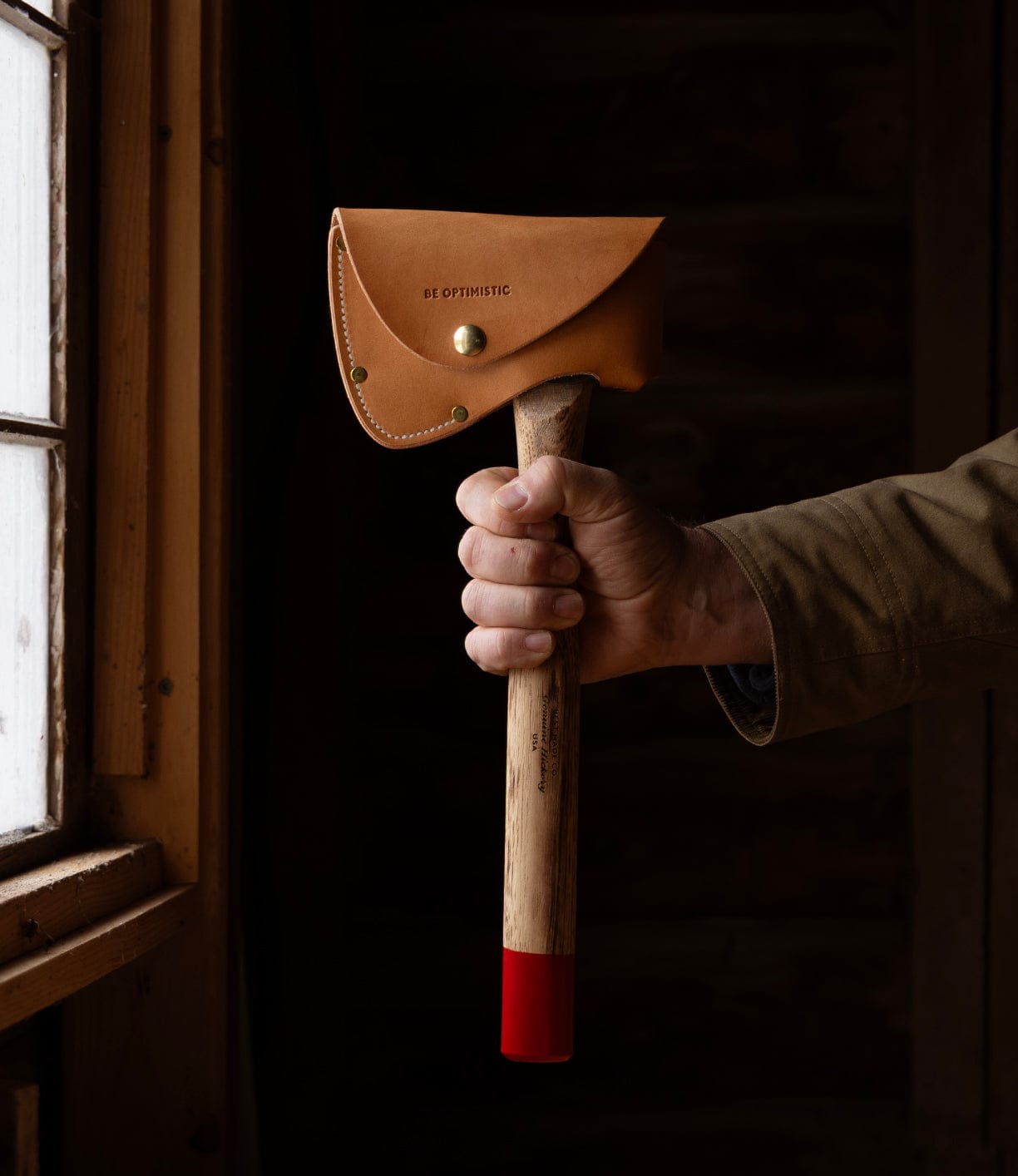 A person in a brown jacket holds Best Made Co.'s Spark (Straight-Hold Edition), featuring an Appalachian hickory handle and a red-tipped high-carbon steel axe head, sheathed in tan leather reading BE OPTIMISTIC. Natural window light fills the scene.
