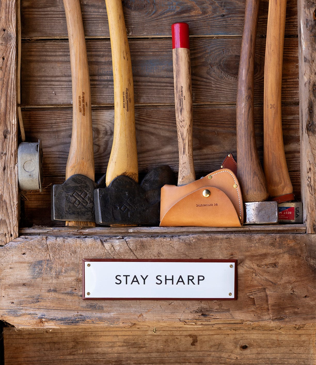 Axes and hammers with wooden handles stand upright by a rustic wall. Below, a white “Stay Sharp” Enamel Steel Sign by Best Made Company Co. displays the bold message in black letters, hand-crafted in Poland.
