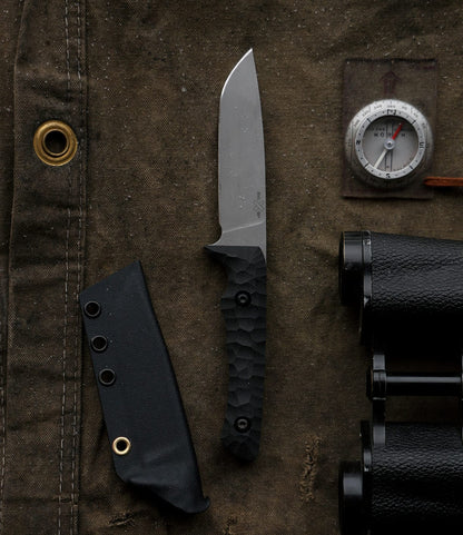 The NWBW Field Knife (BMC Edition) by Best Made Co. has a black textured handle and lies on brown fabric beside its sheath, a compass, and part of black binoculars. A brass grommet is also seen on the fabric.