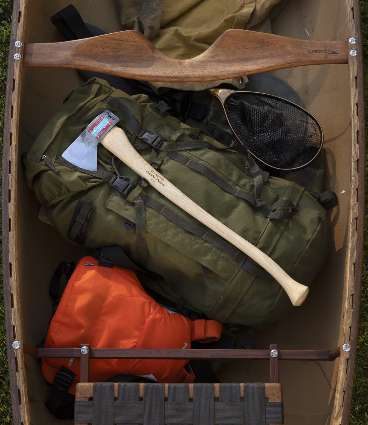 A top view of The Optimist canoe by Best Made Co., packed with camping essentials—a green backpack with a 5160 carbon steel Hudson Bay axe, an orange life jacket, and a fishing net.