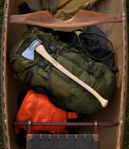 A top view of The Optimist canoe by Best Made Co., packed with camping essentials—a green backpack with a 5160 carbon steel Hudson Bay axe, an orange life jacket, and a fishing net.