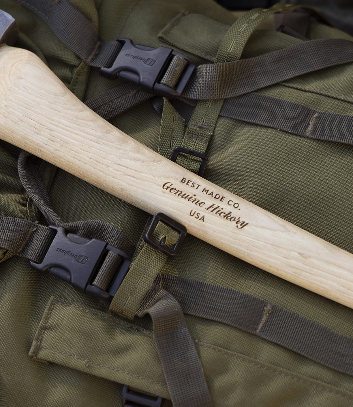 A close-up of The Optimist axe by Best Made Co., featuring a wooden handle, resting on a green backpack with straps and black plastic buckles.