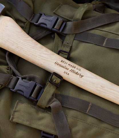 A close-up of The Optimist axe by Best Made Co., featuring a wooden handle, resting on a green backpack with straps and black plastic buckles.