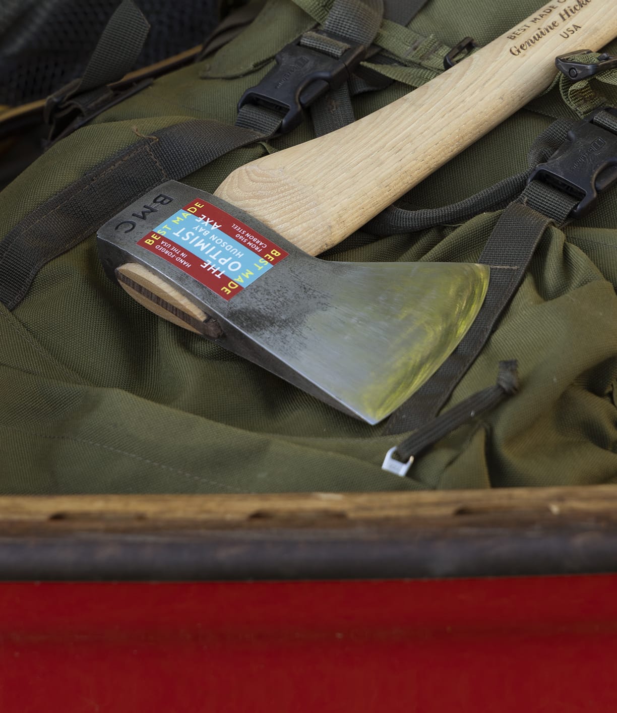 The Optimist by Best Made Co., featuring a wooden handle and colorful label, rests on a green backpack with black straps in a red container. Its head is forged from American 5160 carbon steel.