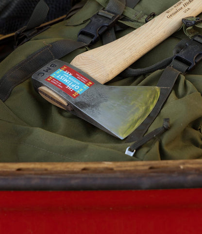 The Optimist by Best Made Co., featuring a wooden handle and colorful label, rests on a green backpack with black straps in a red container. Its head is forged from American 5160 carbon steel.