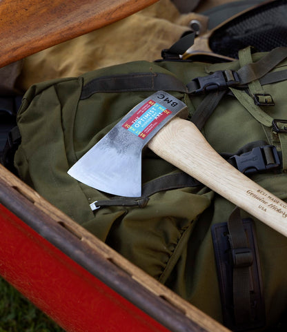 The Optimist axe by Best Made Co. with a wooden handle and sticker sits on a green backpack inside a red canoe, surrounded by outdoor gear—ideal for the prepared canoeist's next camping adventure.