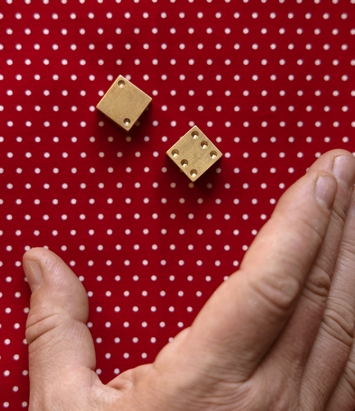 A hand prepares to roll two Solid Brass Dice by Best Made Co., crafted from solid brass, on a red surface with white polka dots.