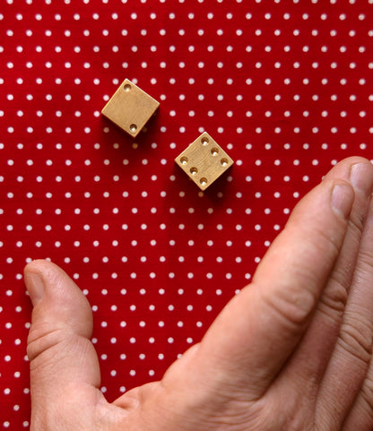 A hand prepares to roll two Solid Brass Dice by Best Made Co., crafted from solid brass, on a red surface with white polka dots.