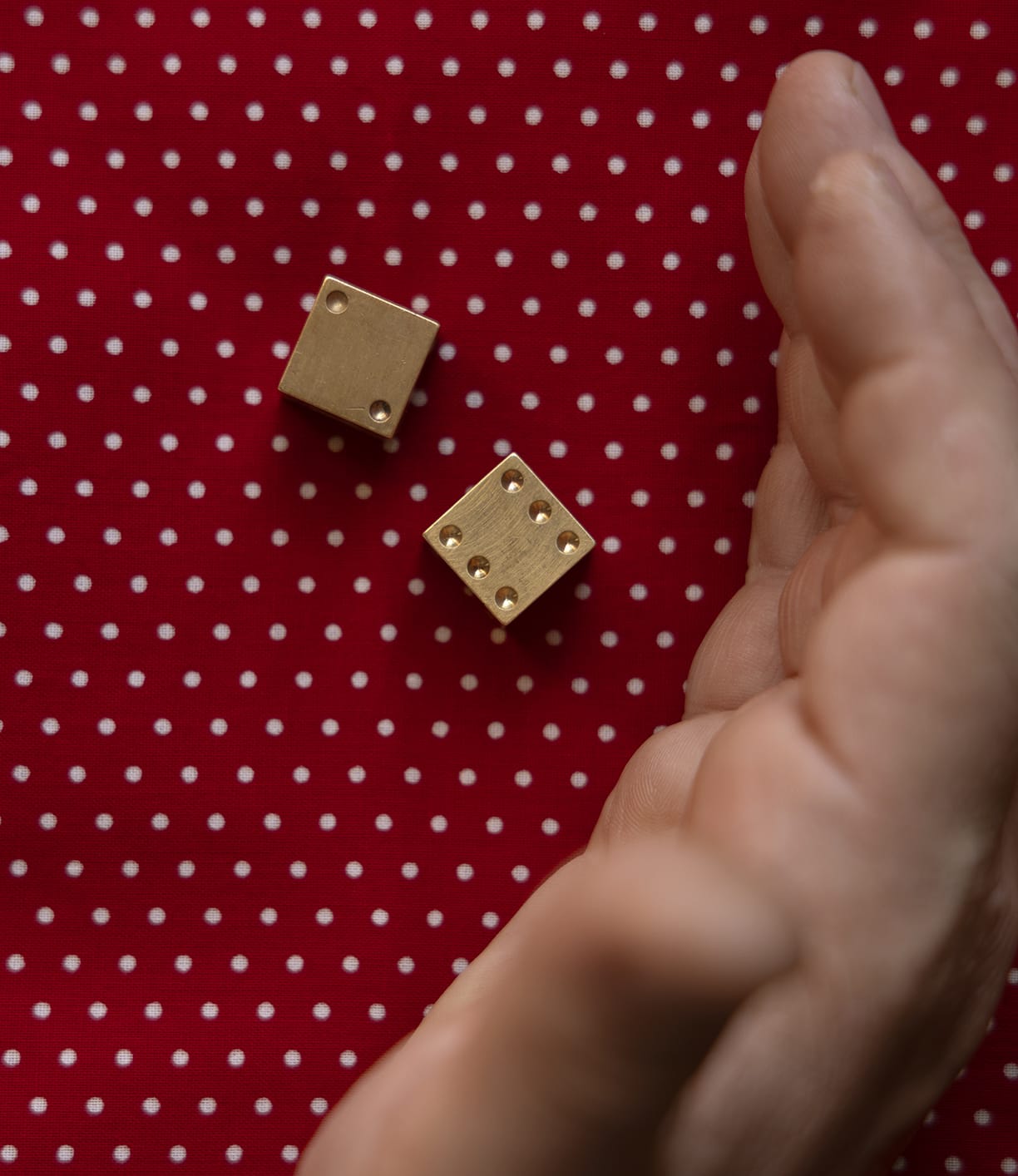 A hand is cupped near two Solid Brass Dice from Best Made Co. on a red surface with white polka dots, showing the numbers one and five.