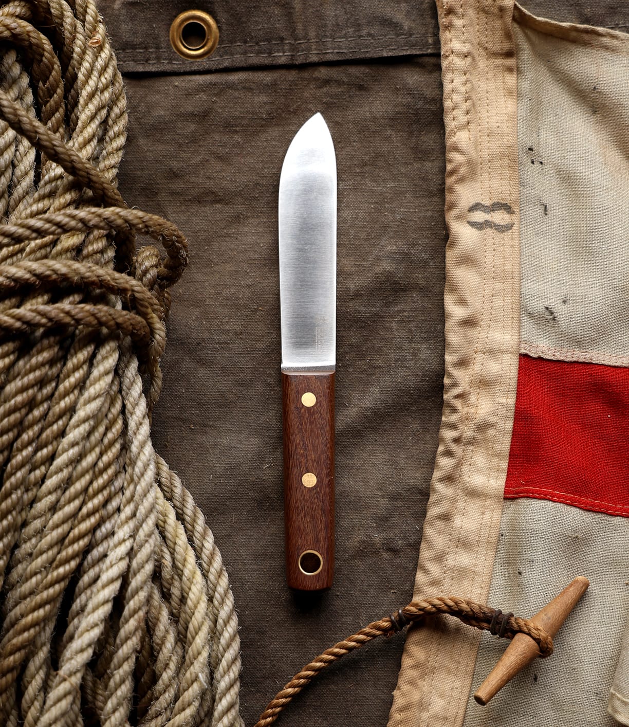 A Best Made Co. Sailor’s Knife with a Brazilian Teak handle rests on brown fabric between coiled rope and canvas with a red stripe.