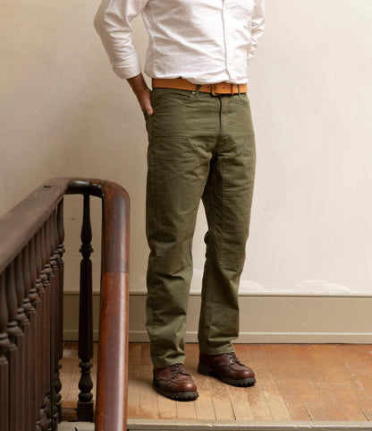 Wearing a white long-sleeve shirt and Best Made Co. Field Pant Special Edition Olive Drab, paired with a brown belt and shoes, a person stands on a wooden floor beside a staircase railing with hands in their pockets.
